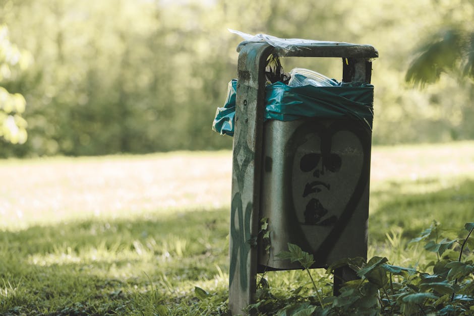 A weathered metal rubbish bin located outdoors on a grassy area, with the top partially open revealing a blue plastic bag and other waste inside. The bin has graffiti artwork painted on its front, depicting a stylized face with dark, hollow eyes and an open mouth, giving it an urban, artistic appearance. The surface of the bin shows signs of rust and dirt, indicative of exposure to the elements. Surrounding the bin are green leaves and small plants, and the background features a blurred natural environment with trees and sunlight filtering through the foliage. The scene highlights an irregular waste disposal site, relevant to private rubbish collection services like Rubbish Clearance Barnes, showcasing the importance of proper rubbish management and alternative disposal methods outside typical municipal collection areas.