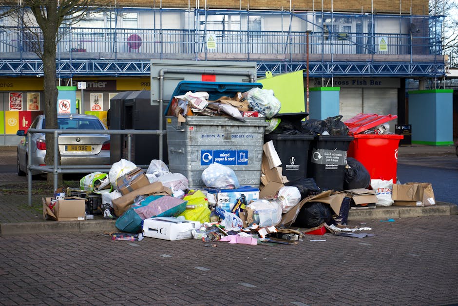 A collection of overflowing rubbish and waste materials gathered around a large grey mixed paper and cardboard recycling bin situated on a paved urban street. The bin, positioned in the foreground, is partially open with assorted paper, cardboard, and plastic packaging spilling out onto the ground, surrounding the base of the bin. Various black and red rubbish bags are piled nearby, some torn or displaced, revealing contents such as plastic bottles, wrappers, and discarded materials. A silver car is parked to the left, adjacent to a metal railing, and behind the refuse are retail units or commercial storefronts with brightly coloured signage and window displays. In the background, a building under renovation is visible, partially covered with blue scaffolding and safety mesh, suggesting recent or ongoing construction activity. The scene reflects an area experiencing issues with waste clearance, common in private waste handling or alternative rubbish collection services, with an emphasis on unmanaged refuse accumulation in an urban environment.