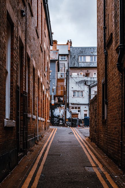 A narrow urban alleyway situated between two brick buildings, with the foreground showing pavement marked by double yellow lines along the edges. The brick walls on either side are reddish-brown with visible mortar joints and occasional black drain pipes running vertically. In the background, there is a small area of open sky with a cloudy, overcast appearance. The alley ends at a cluster of multi-story buildings with variously coloured exterior walls, including grey and white, featuring different window styles and small external staircases. Around the end of the alley, there are black wheelie bins and miscellaneous debris, suggesting prior rubbish clearance activities. The scene appears wet, possibly from recent rain, and shows urban clutter typical of a location where private waste collection or on-site clearance might be required, aligning with independent rubbish removal services such as Rubbish Clearance Barnes.