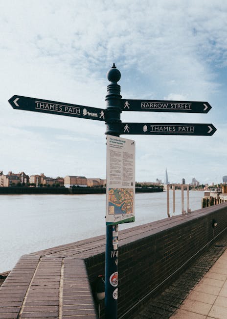 A black metal signpost with four directional arrows stands along a paved walkway beside the river, with a backdrop of a city skyline under a partly cloudy sky. The arrows point to 'Thames Path' and 'Narrow Street,' indicating pedestrian routes along the Thames River in Barnes. The signpost features a spherical finial at the top and a rectangular informational plaque mounted below, which displays a map and text. To the right of the sign, there is a black wooden bench with a slatted seat and backrest, positioned close to the water's edge with remaining space on the paved walkway. The river in the background reflects the sky and distant buildings, and the scene suggests a peaceful riverside setting suitable for walking and local sightseeing, possibly within a reasonable distance of independent waste collection or rubbish clearance services operating in the area, as part of private waste handling or on-site clearance activities. The overall environment appears bright with natural daylight, and the landscape emphasizes the urban riverside atmosphere without additional clutter or objects in the immediate vicinity.