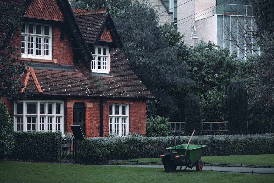 In the foreground of the image, a green wheelbarrow with black handles and red wheels is positioned on a paved pathway, filled with garden waste including sticks and debris. Near the wheelbarrow, there is a black trash bag, possibly containing additional garden refuse. The middle ground features a well-maintained section of a garden bordered by low, decorative iron fencing and neatly trimmed hedges. The background showcases a traditional red-brick house with a dark, sloped tiled roof, multiple white-framed sash windows, and a black front door. Surrounding the house are lush green trees and shrubbery, creating a natural, leafy environment. Modern high-rise buildings are visible further behind, contrasting with the traditional architecture of the house. The overall scene appears to be part of a private garden or outdoor space where garden waste is being collected or prepared for disposal, aligning with independent rubbish removal or clearance services like those offered by Rubbish Clearance Barnes, and demonstrating a typical garden tidy-up in an urban setting.