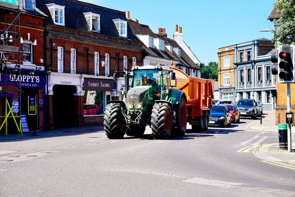 A green and black tractor with large front and rear tires is parked on an urban street, attached to an orange waste collection container at the rear. The scene is set in a town center with a row of brick buildings on the left side, featuring shopfronts including a sports bar and a salon, with signage visible. The street is paved with asphalt, with several parked cars lining the curb on the right side. A traffic light with red signal is visible on the right, along with a small green post box near the curb. The sky is clear and blue, and there are trees visible in the background, indicating a bright, sunny day. This scene reflects private waste handling, possibly as part of an alternative rubbish removal service by Rubbish Clearance Barnes, serving local businesses in the area of High Street Barnes.